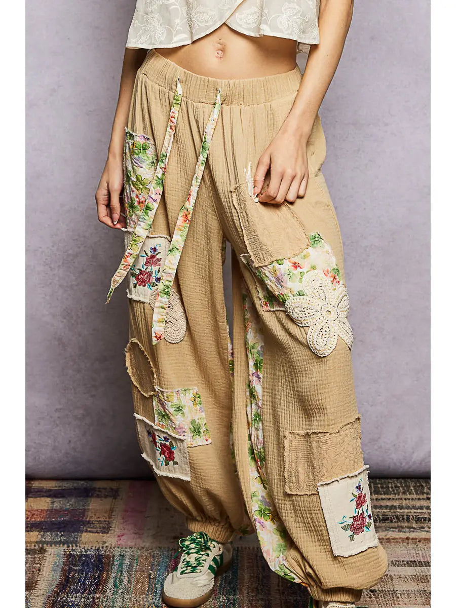 Person wearing beige patchwork pants with floral and lace details on a textured floor.