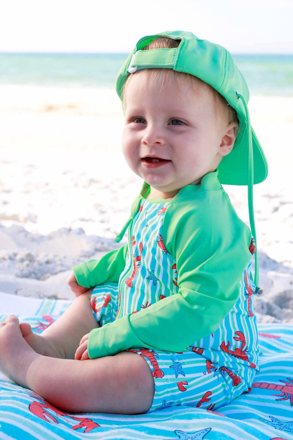 Baby in green swimsuit and cap sitting on a beach towel with ocean view