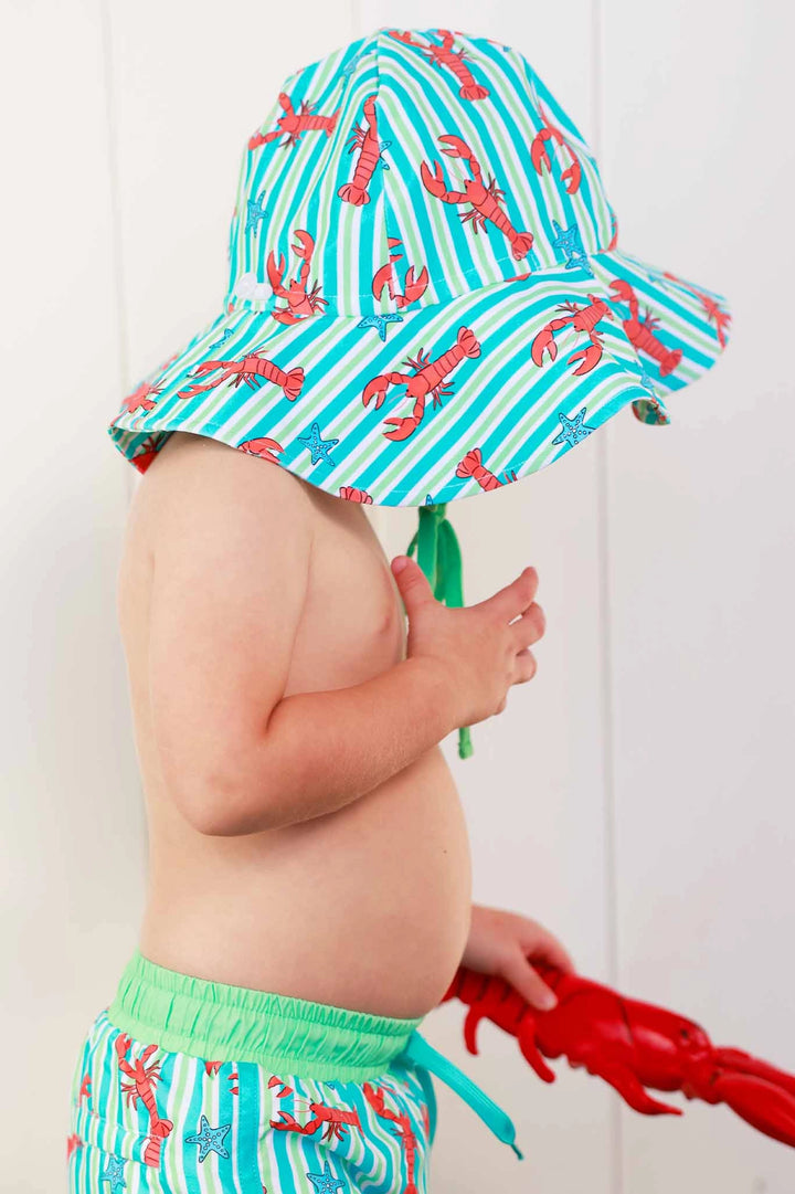Child wearing a colorful lobster-patterned hat and swim trunks against a white background