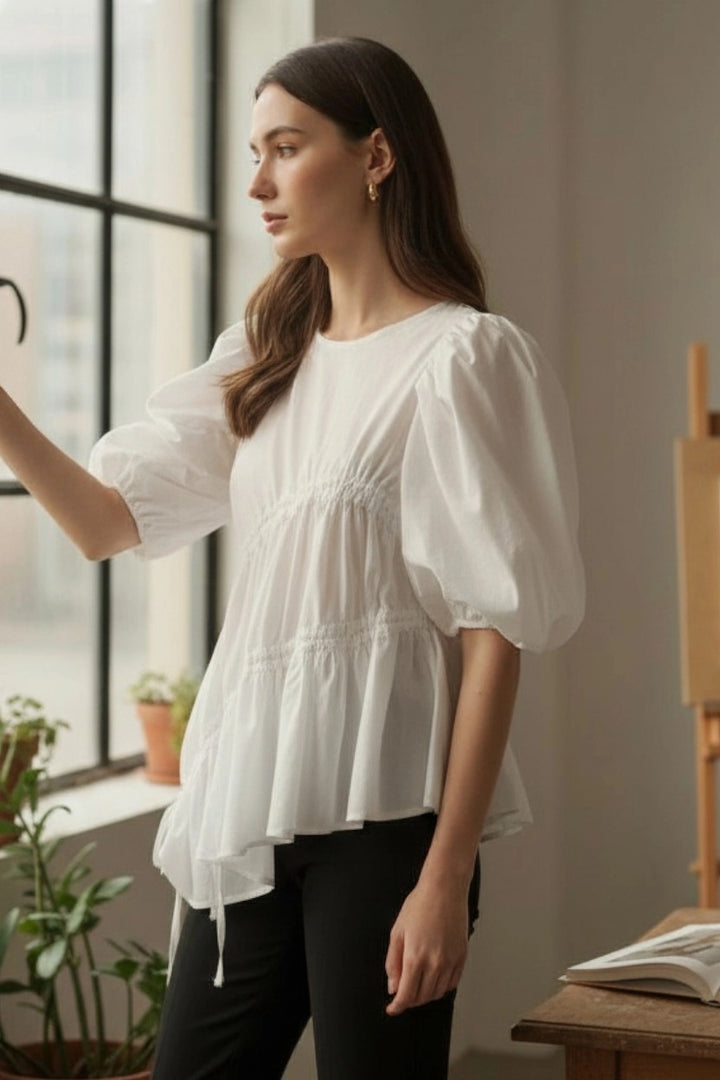 Woman wearing a white blouse standing by a window with plants around.