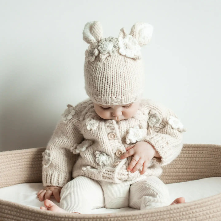 Baby in the natural Blossom Baby Cardigan and matching Blossom Bunny Baby Hat sitting in a woven basket