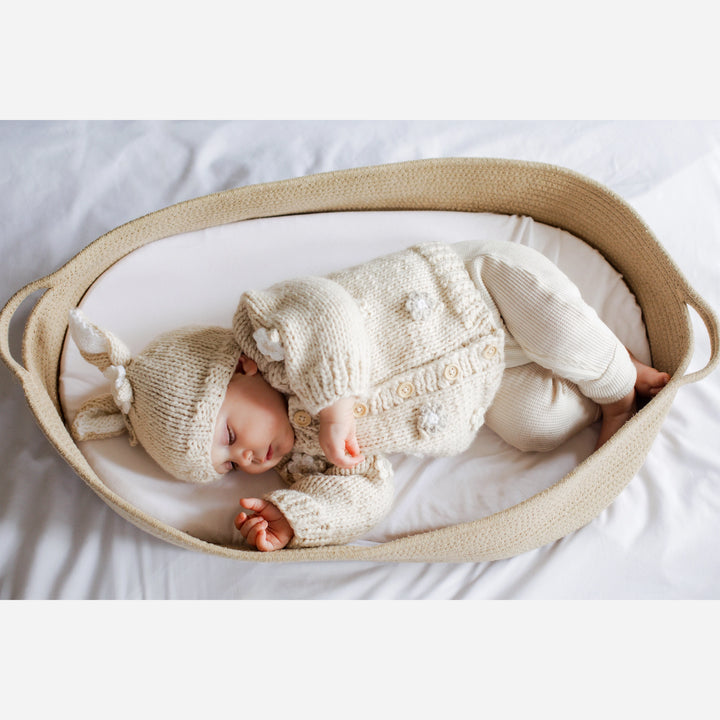 Sleeping baby in a woven basket wearing the Blossom Baby Cardigan and matching bunny hat