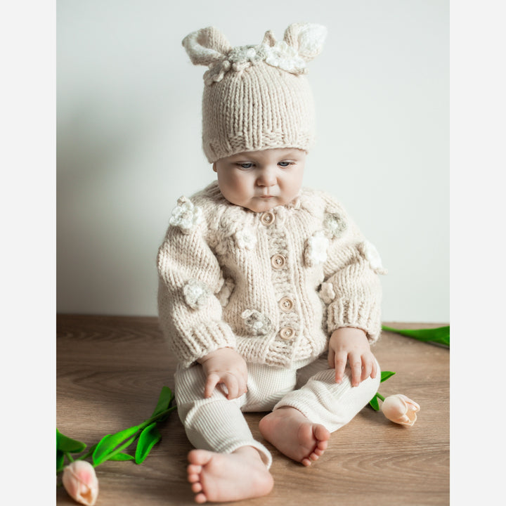Baby in the Blossom Baby Cardigan and Blossom Bunny Baby Hat sitting on a wood floor with tulips