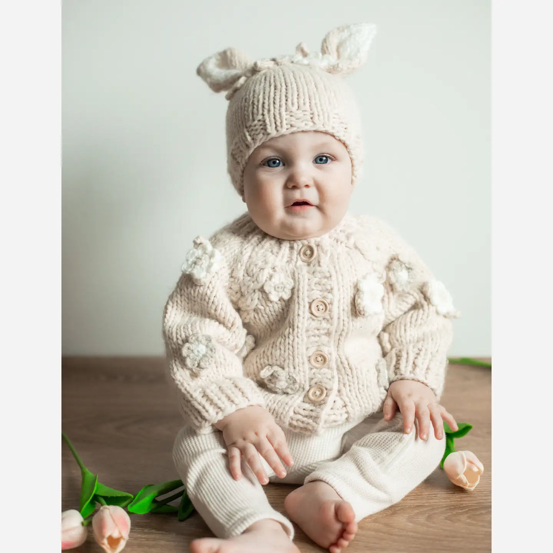 Baby sitting in the Blossom Baby Cardigan with crocheted flowers and matching bunny ear hat