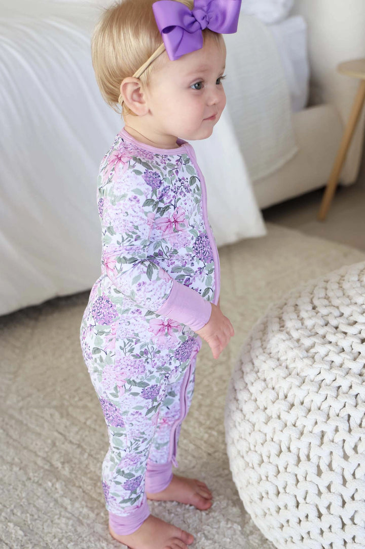 Baby in a floral onesie with a purple bow standing on a carpeted floor.