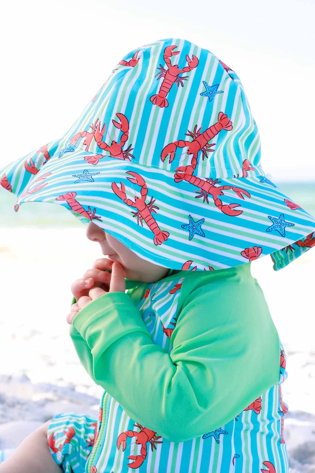 Child wearing a colorful sun hat with lobster design on a beach.