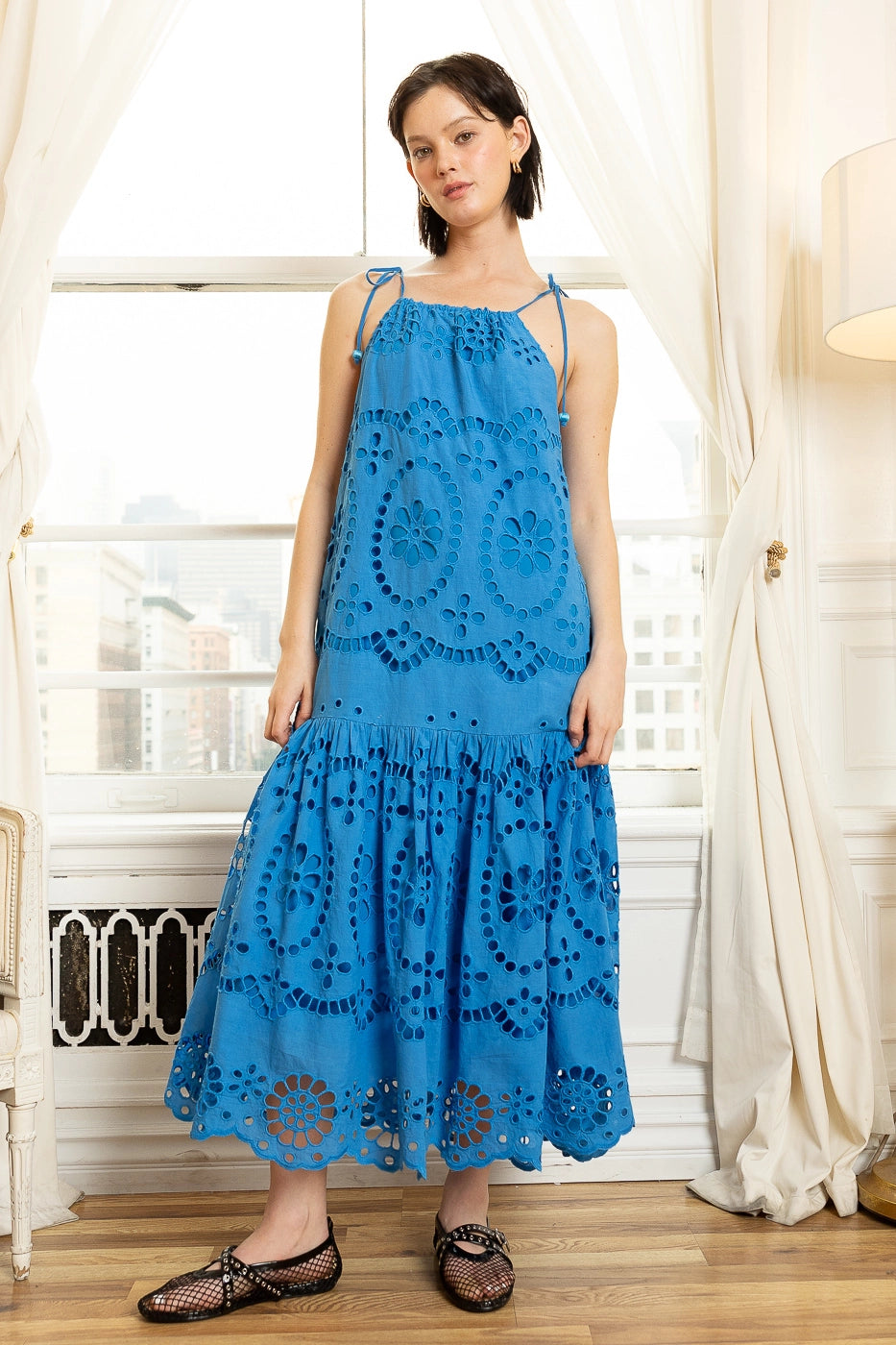 Woman wearing a blue lace dress standing indoors with white curtains and a radiator in the background.