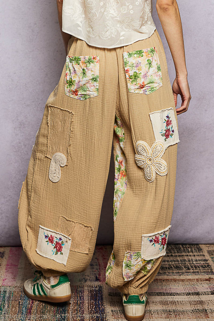 Beige pants with floral embroidery and patches on a textured floor.