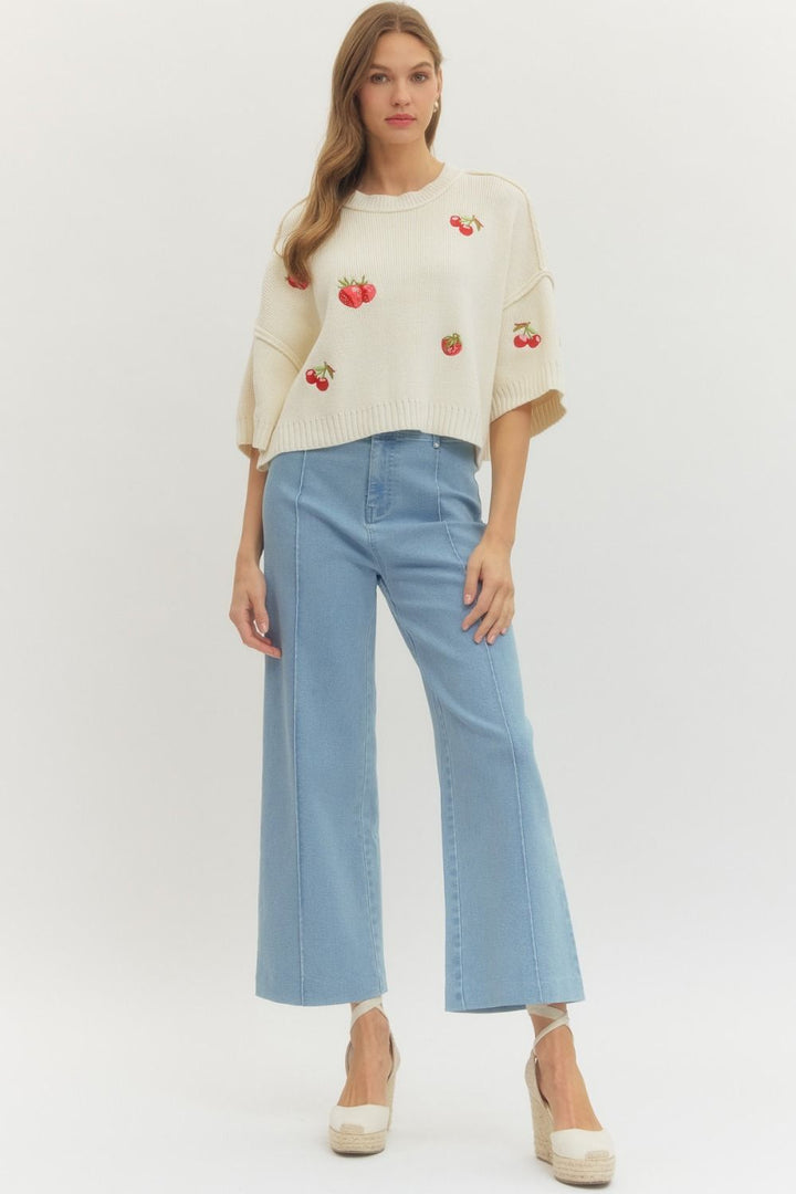 Woman wearing a cream sweater with red apple pattern and light blue jeans on a white background