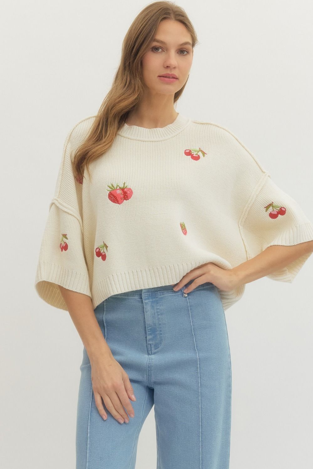 Woman wearing a cream sweater with cherry embroidery and light blue jeans on a white background