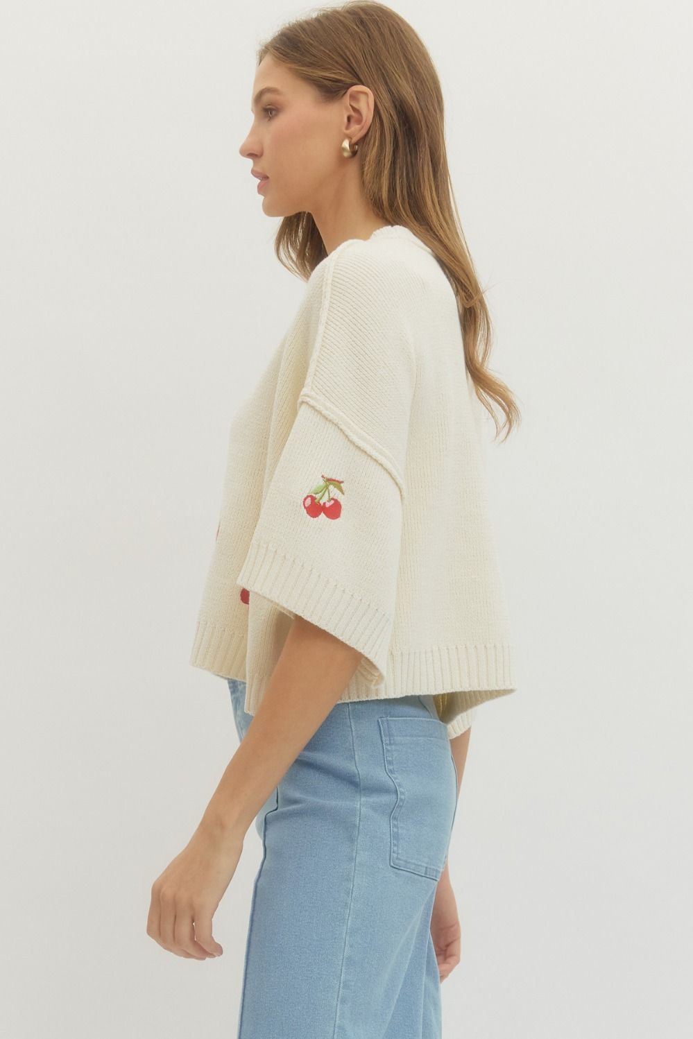 Woman wearing a cream-colored sweater with cherry design and light blue jeans on a white background