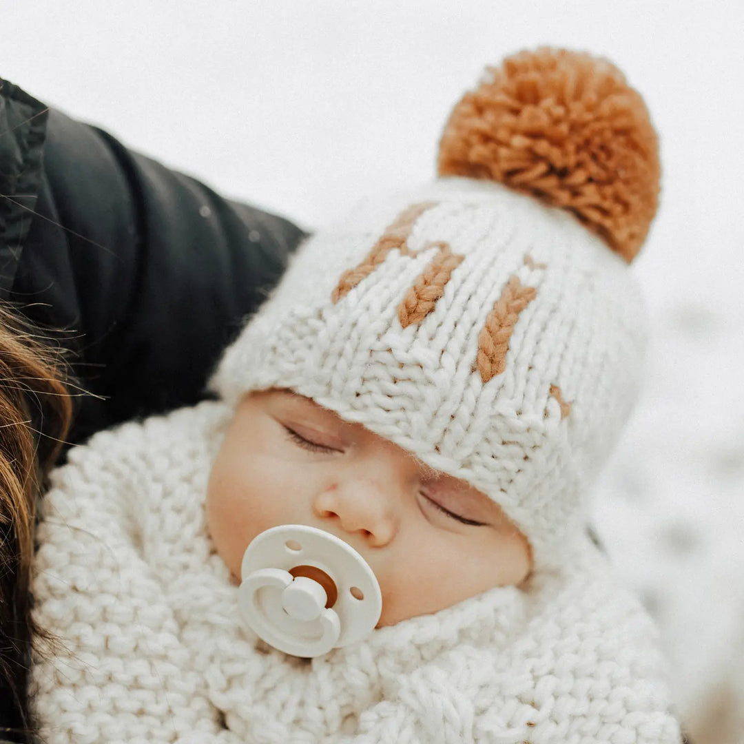 Close-up of a baby with a pacifier wearing the natural Hi There Baby Hat with pecan pom pom