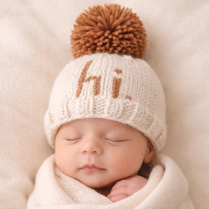 Sleeping newborn wearing the natural Hi There Baby Hat with pecan hi. lettering and pom pom