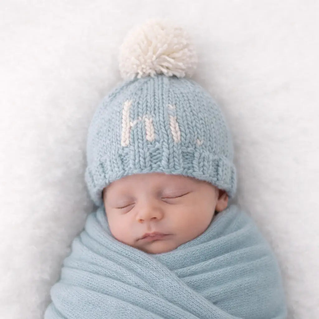 Swaddled newborn wearing the surf blue Hi There Baby Hat with natural pom pom