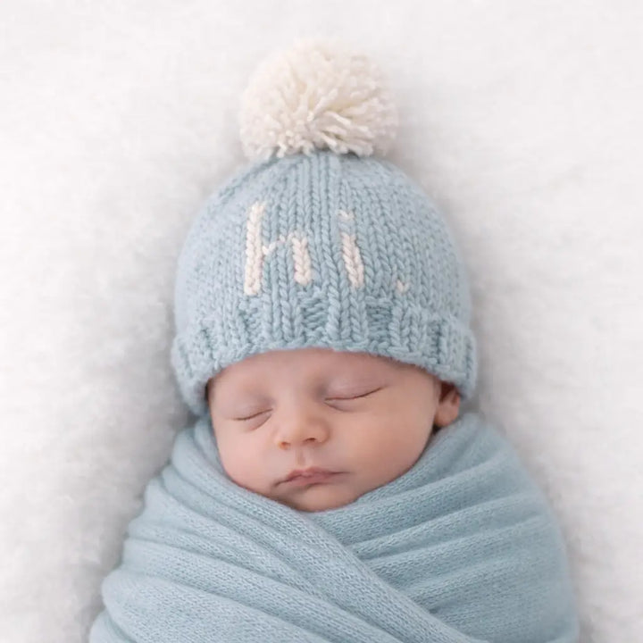 Swaddled newborn wearing the surf blue Hi There Baby Hat with natural pom pom