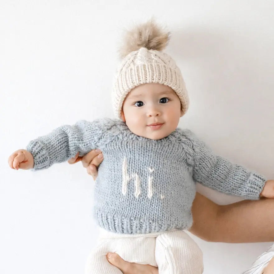 Baby wearing the surf blue Hi There Baby Sweater with natural hi. lettering and a pom pom beanie