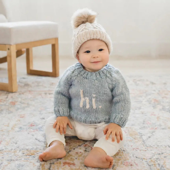 Smiling baby sitting on a rug in the surf blue Hi There Baby Sweater