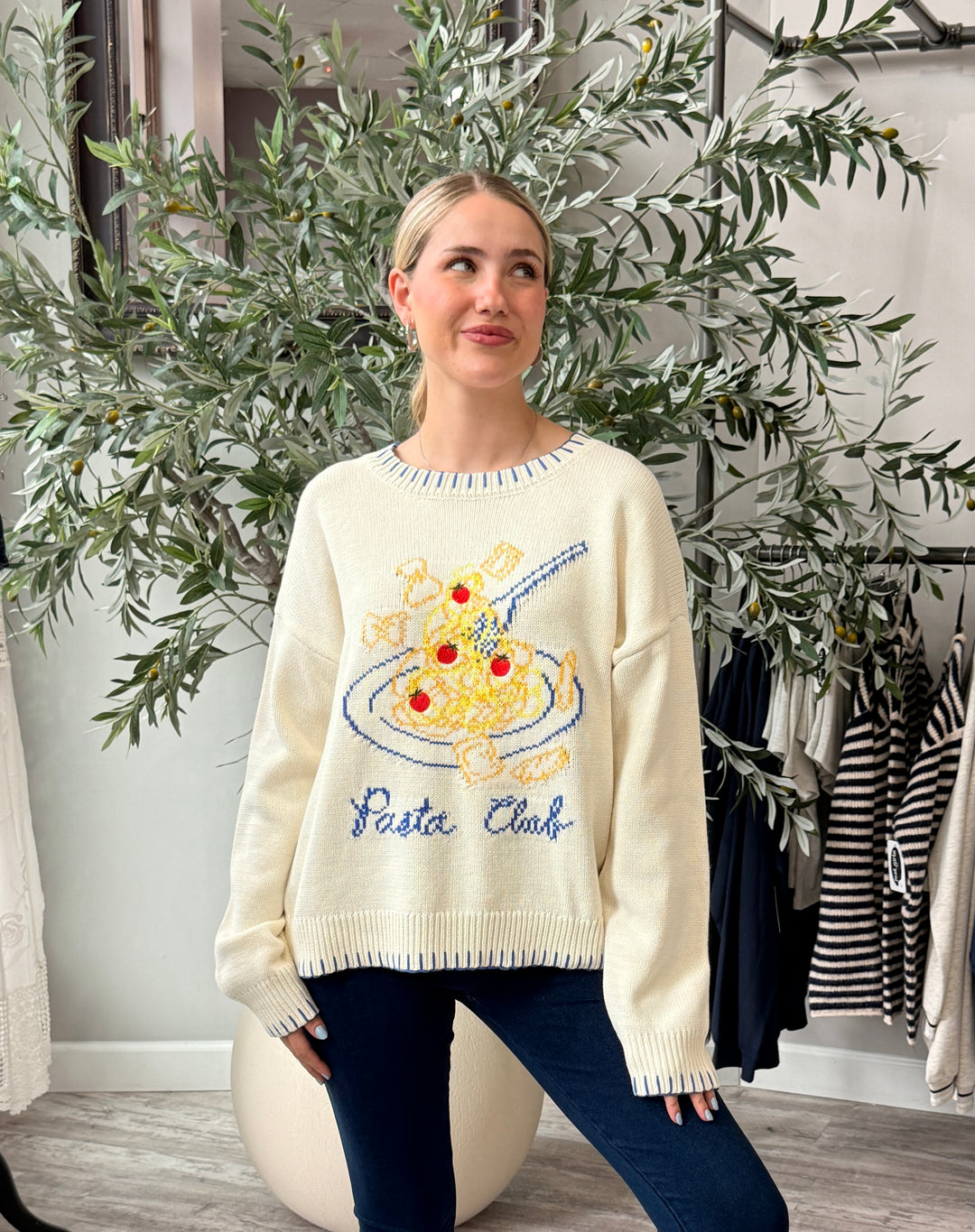 A woman wearing a cream-colored knit sweater with a colorful spaghetti and pizza graphic and the words 'Pasta Club' embroidered on the front, paired with dark blue jeans, posed in front of a potted olive tree in a boutique setting.