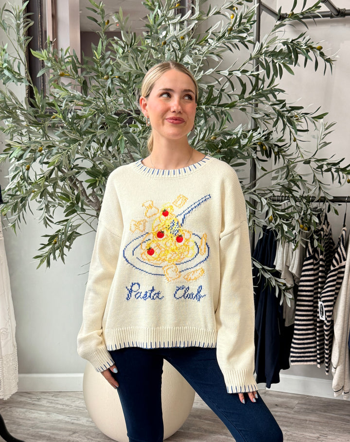A woman wearing a cream-colored knit sweater with a colorful spaghetti and pizza graphic and the words &