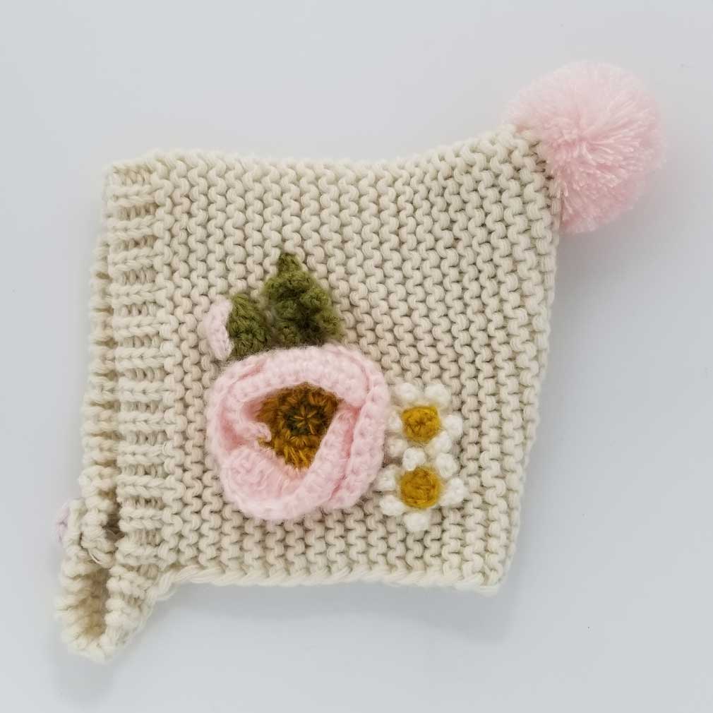 Close-up of a natural knit baby bonnet with a pink mohair poppy, green leaves and pink pom pom