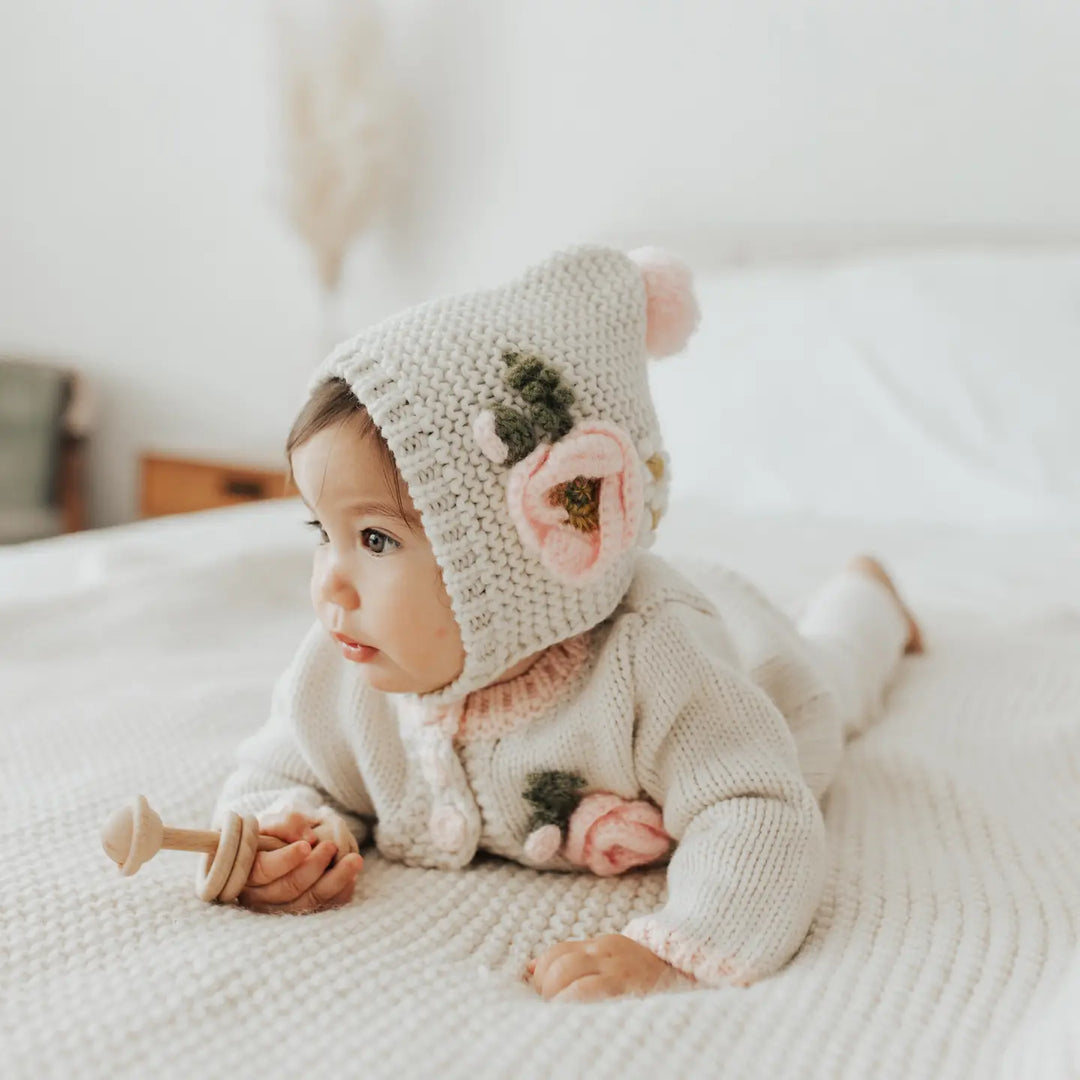 Baby lying on a bed wearing the natural Poppy Bloom Baby Cardigan with pink mohair poppy blossoms