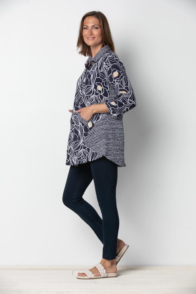 A woman is standing against a plain background, wearing a floral tunic top and the 'Keep Up Easy Pant' in denim. She has accessorized with earrings, a bracelet, and is wearing open-toe flat sandals.